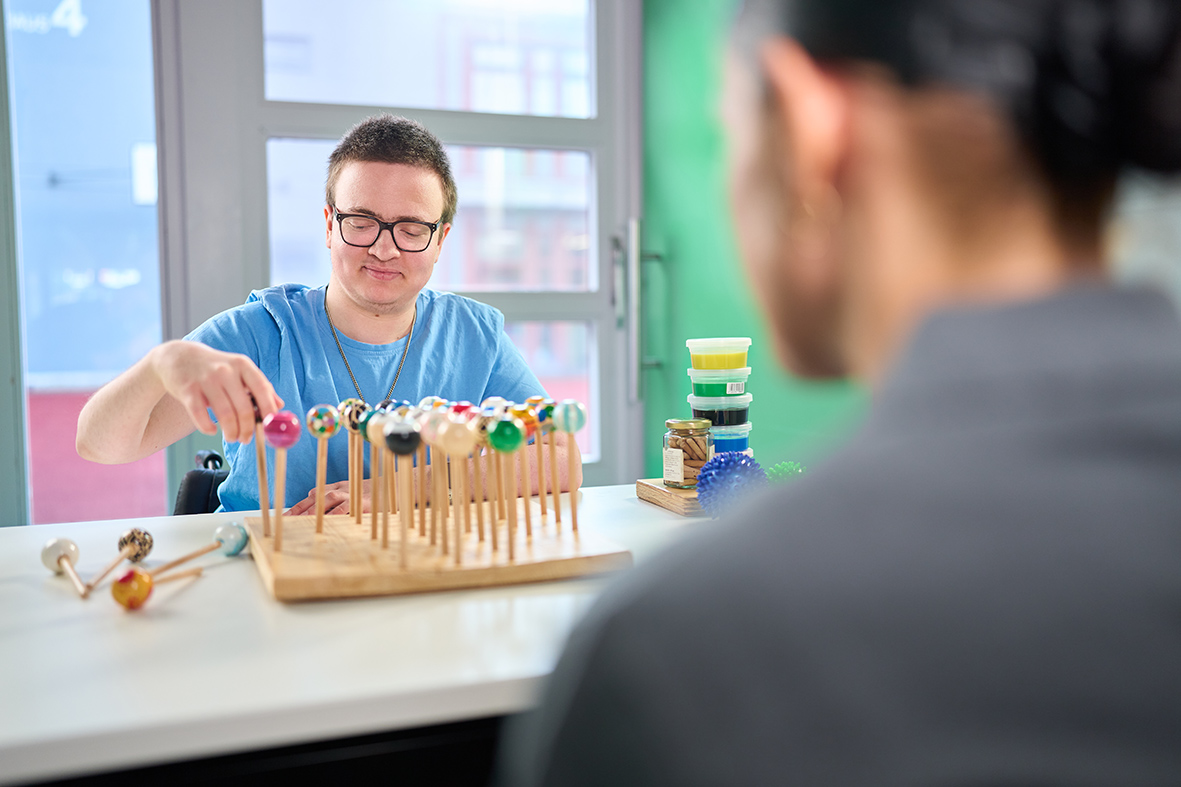 Auszubildender macht Übung in der Ergotherapie Ein Mann mit Brille und blauem Kittel montiert bunte Kugeln auf Zahnmodelle, ein anderer beobachtet. Werkzeug und Materialien liegen auf dem Tisch.