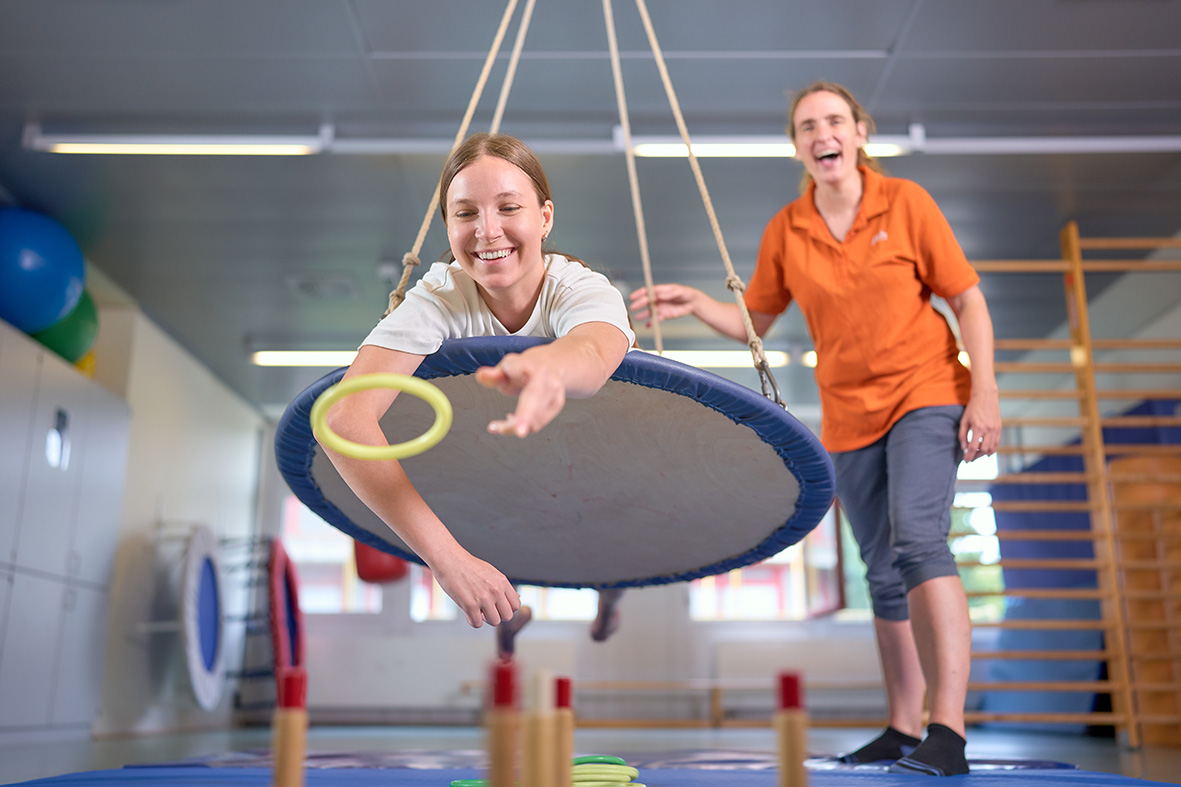 Zwei Personen in einer Trampolinhalle: Eine schwebt mit Ring über einem Mini-Trampolin, die andere beobachtet lachend.