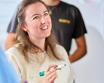 Eine Frau schreibt etwas auf ein Whiteboard Eine Frau hält einen Stift und lächelt, im Hintergrund steht ein Mann in schwarzem Shirt. Beide wirken in einem hellen Raum.
