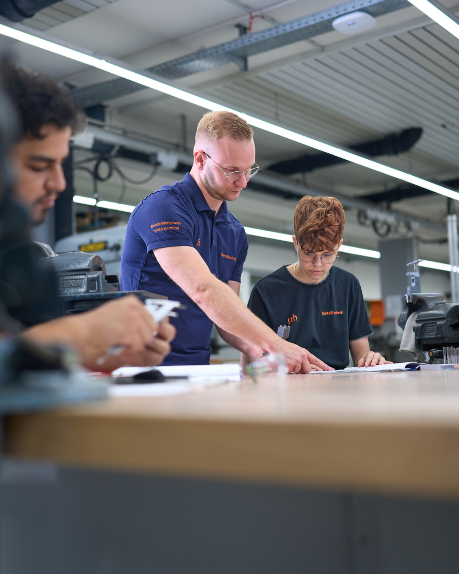 Drei Personen arbeiten in einer Werkstatt an Motorenkomponenten, zwei stehen und beobachten, eine arbeitet am Tisch.
