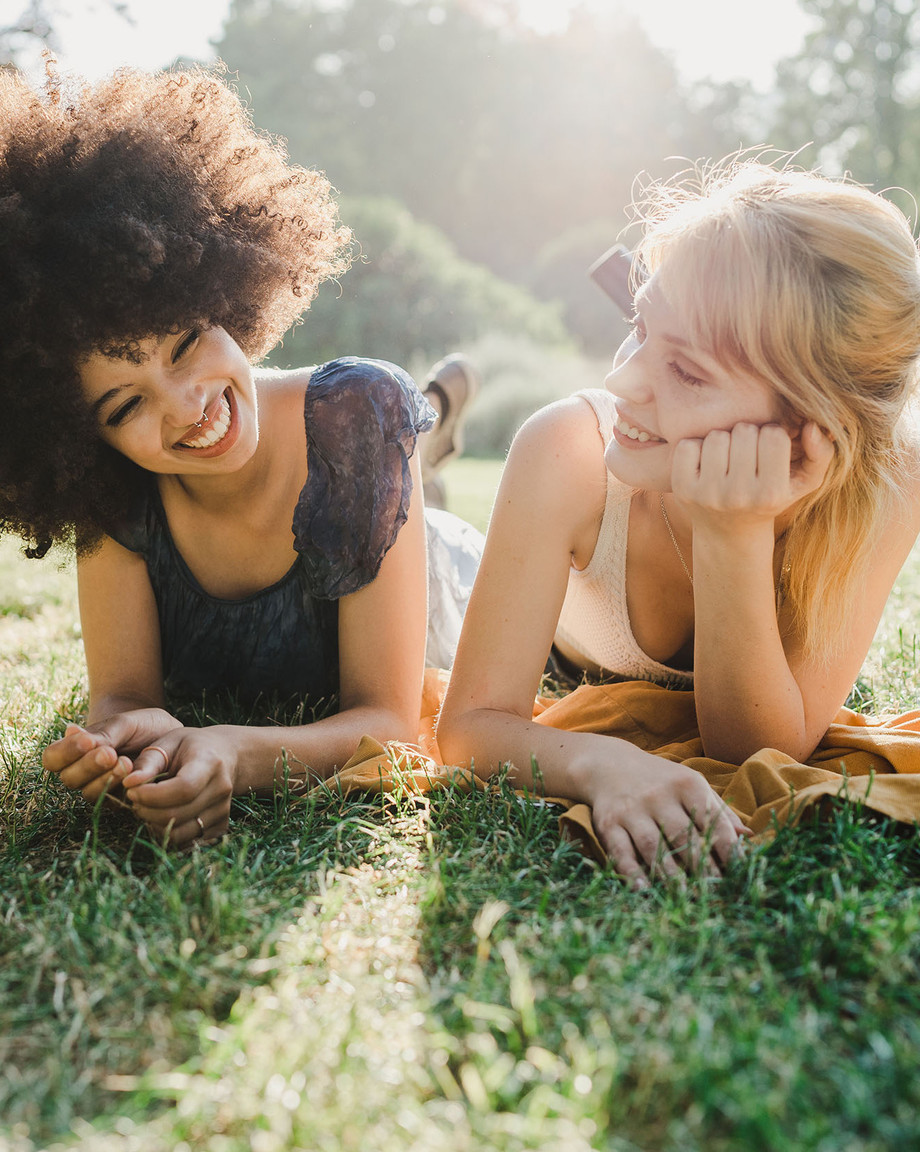Zwei junge Frauen liegen nebeneinander auf einer Wiese und unterhalten sich glücklich miteinander.