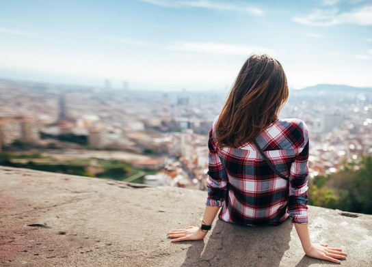 Eine junge Frau sitzt auf einer Mauer und schaut auf eine Stadt im Ausland.