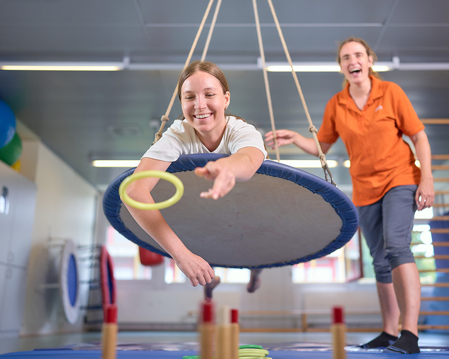 Zwei Personen in einer Trampolinhalle: Eine schwebt mit Ring über einem Mini-Trampolin, die andere beobachtet lachend.