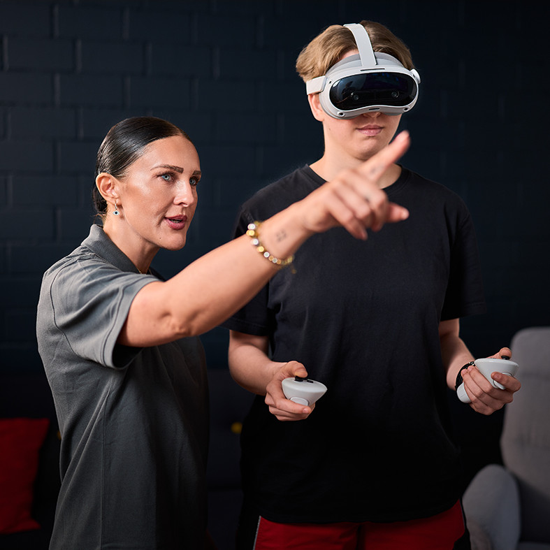 Eine Person erklärt einer anderen mit VR-Brille und Handcontroller eine Anwendung. Beide stehen in einem Raum mit dunkler Wand.