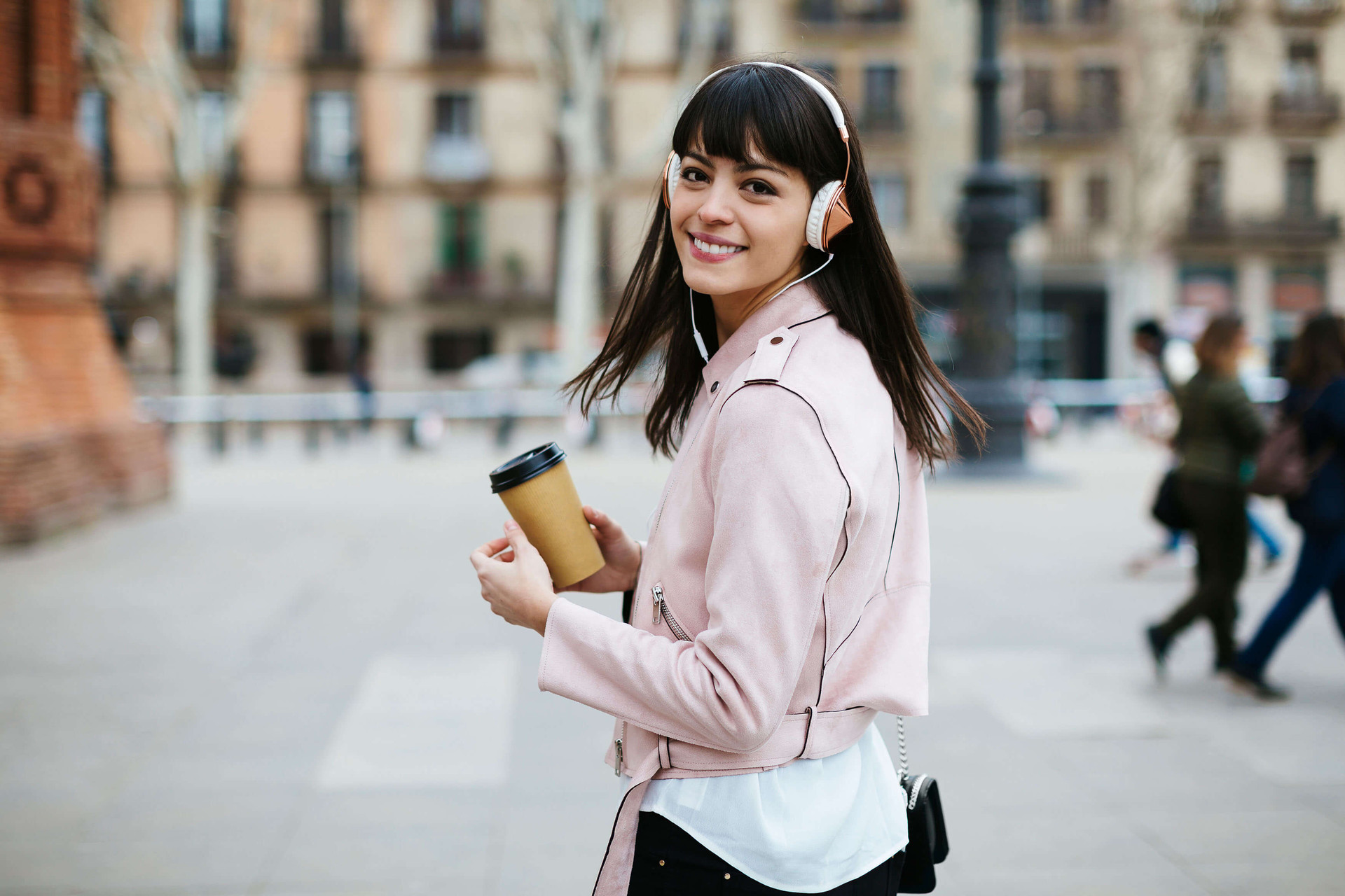 Junge Frau mit Kopfhörern läuft in der Stadt mit Kaffee in der Hand und schaut zurück.