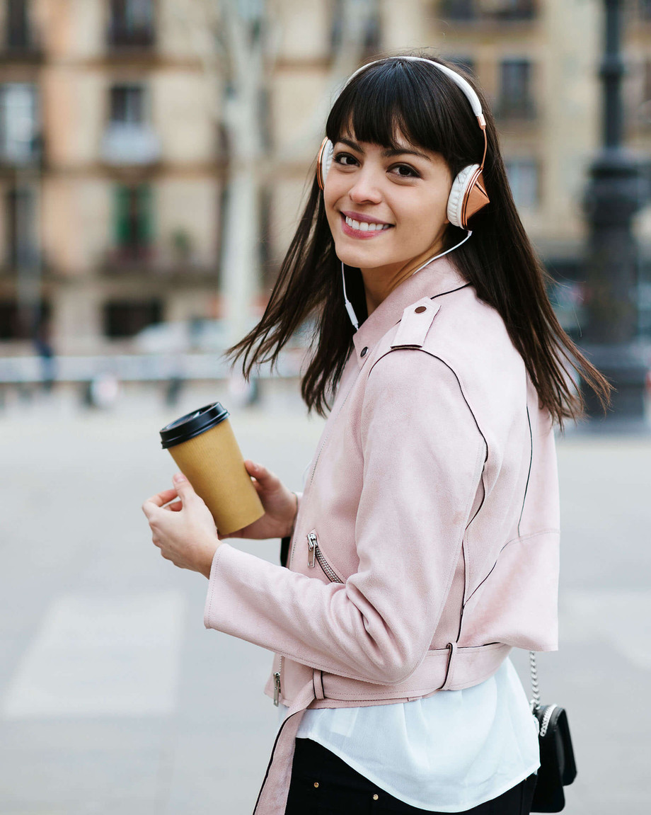 Junge Frau mit Kopfhörern läuft in der Stadt mit Kaffee in der Hand und schaut zurück.