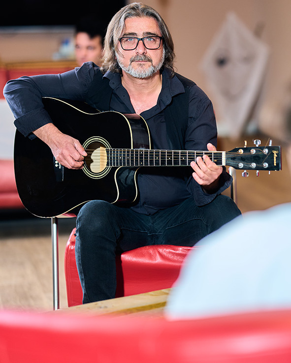 Ein Mann sitzt und spielt Gitarre, im Hintergrund weitere Personen.