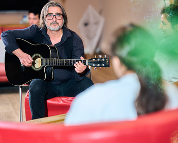 Ein Mann sitzt und spielt Gitarre, im Hintergrund weitere Personen.