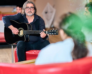 Ein Mann sitzt und spielt Gitarre, im Hintergrund weitere Personen.