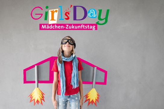 Junges Mädchen mit Fliegerbrille hat Flugzeugflügel um und schaut auf das Girls’ Day Logo hoch.