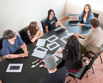Gruppe sitzt am Tisch und lernt etwas Eine Gruppe von sieben Personen sitzt im Kreis an einem Tisch mit Papieren und diskutiert. Eine Person arbeitet an einem Laptop.