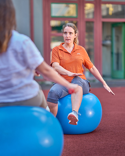 Physiotherapeutin mit Patientin auf Gymnastikball Eine Person sitzt auf einem blauen Gymnastikball, während eine Trainerin eine Übung korrigiert. Beide befinden sich im Freien auf einem Sportplatz.