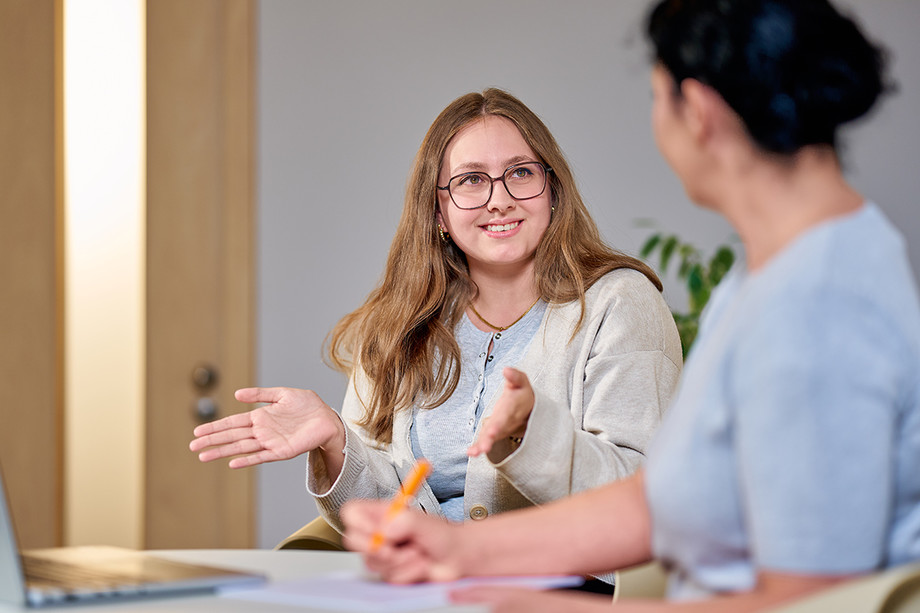 Auszubildende in Therapie Zwei Personen sitzen an einem Tisch, eine Frau erklärt mit Gesten etwas und lächelt, daneben Notizen und Laptop.