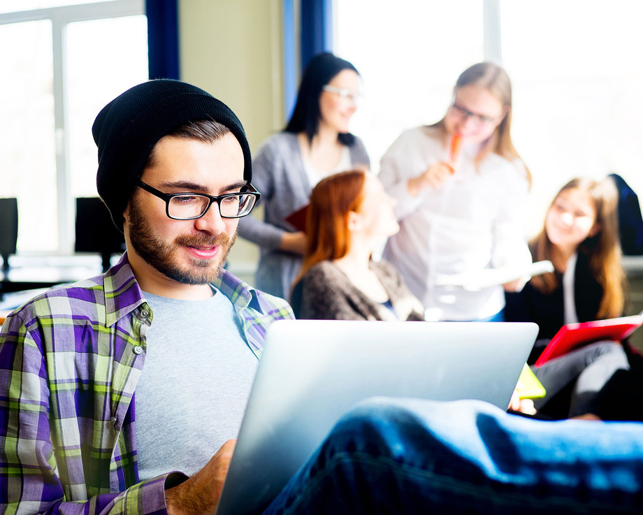 Ein Mann mit Brille und Mütze arbeitet an einem Laptop in einem Büro mit mehreren Personen im Hintergrund.