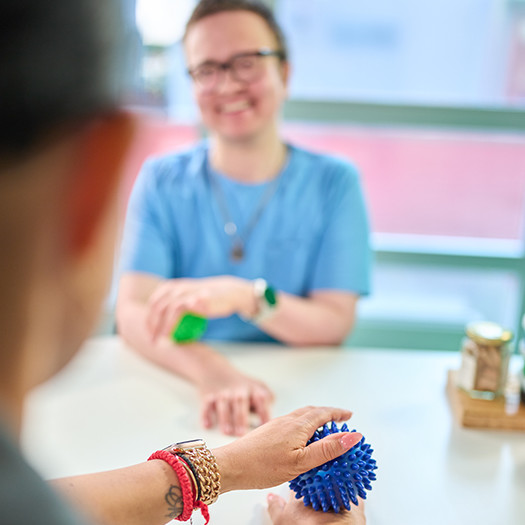 Zwei Personen sitzen an einem Tisch, eine zeigt der anderen ein blaues Massagegerät für die Hand.