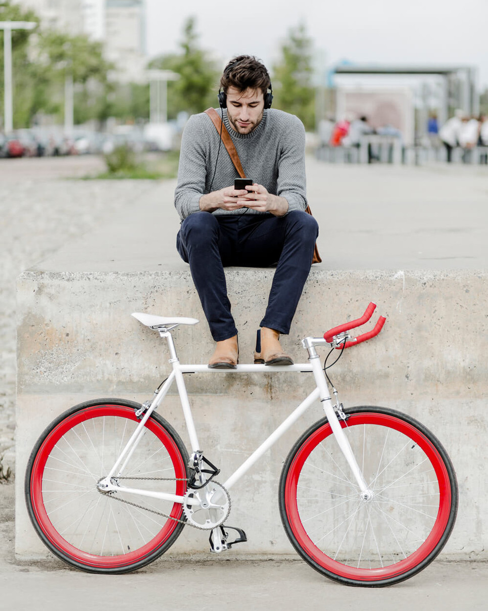 junge Mann mit Fahrrad sitzt auf einer Mauer und schaut auf sein Smartphone.